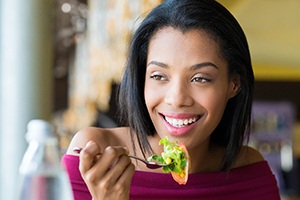A young woman wearing an off-the-shoulder blouse is eating a salad while wearing dental implants in Bedford
