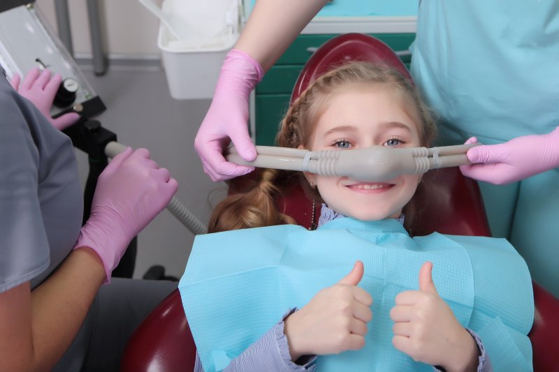 Girl giving thumbs up and smiling as two dentists place nasal mask